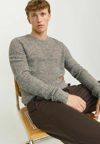 Grijs getextureerd gebreid sweatervest met een ronde hals, gecombineerd met een paar bruine broeken. Het vest heeft lange mouwen en een ontspannen pasvorm en zit op een stoel.