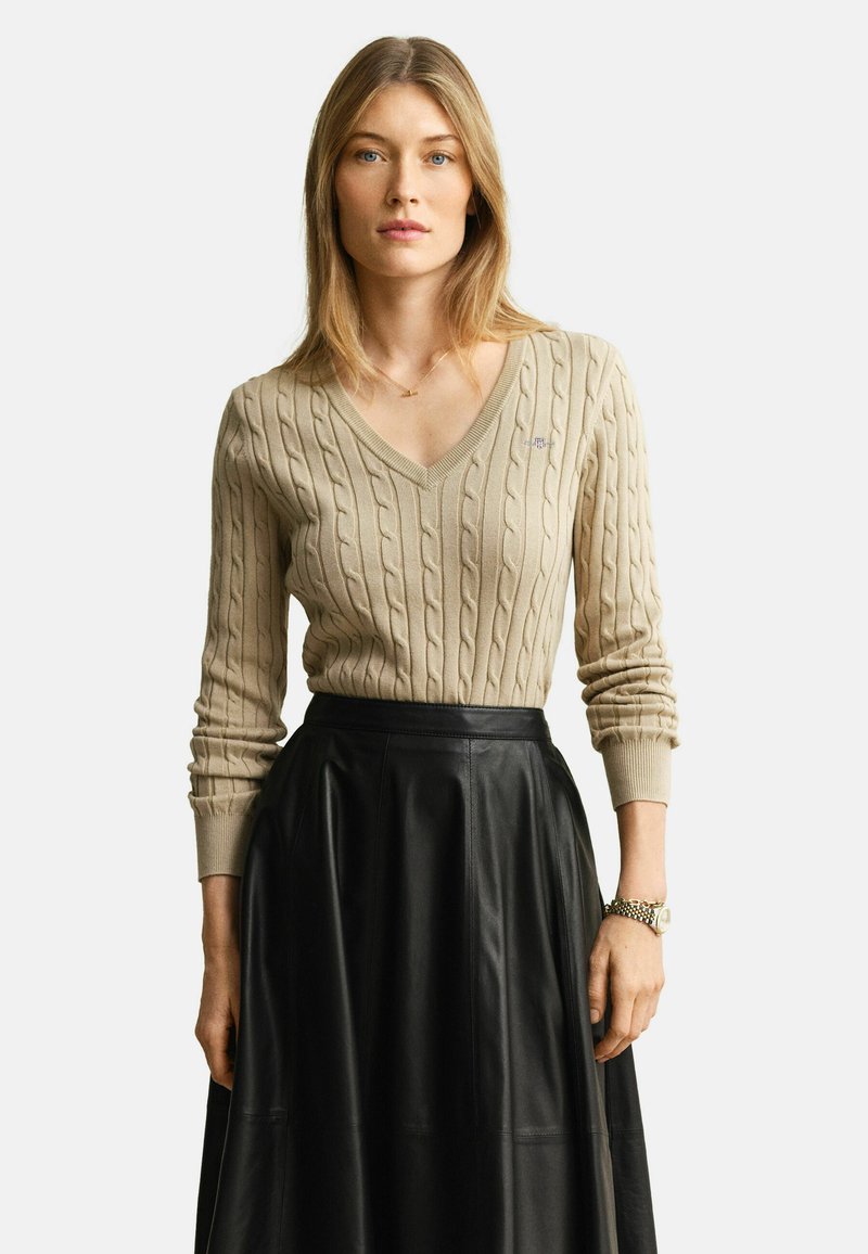 Femme portant un pull beige à col en V en maille torsadée et une jupe en cuir noir taille haute, debout devant un fond blanc uni.