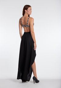 Black high-slit skirt paired with a patterned bralette featuring geometric shapes. Model wears black heeled shoes, showcasing smooth fabric and cut.