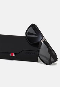 Czarne okulary przeciwsłoneczne Carrera z ciemnymi soczewkami spoczywające na dopasowanym czarnym etui z logo Carrera oraz małą czerwoną, białą i czarną metką z tkaniny.