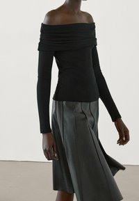 Maglia nera a spalle scoperte con maniche lunghe abbinata a una gonna a pieghe in pelle nera. La maglia presenta un collo drappeggiato e una silhouette aderente.