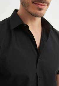 Chemise noire à manches courtes avec col pointu, présentant une texture lisse et une fermeture à boutons à l'avant.