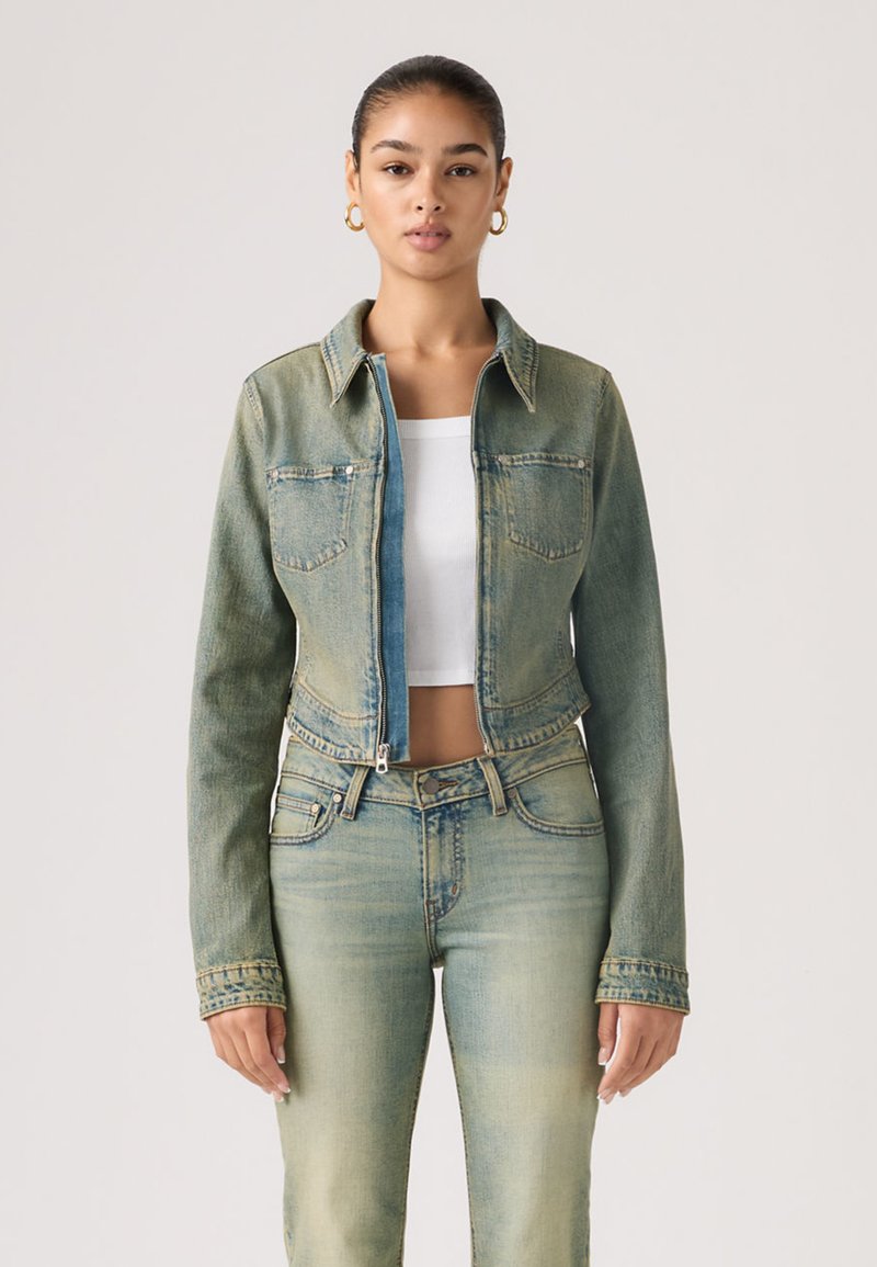 Jeune femme aux cheveux tirés en arrière portant des boucles d'oreilles en créole dorées, une veste en jean verte délavée par-dessus un crop top blanc, et un jean taille haute assorti.