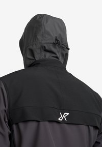 Bakifrån av en person som bär en mörkgrå jacka med huva och en svart yoke-panel med en liten vit logotyp.