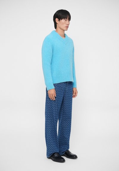 Lichtblauwe, fuzzy v-hals trui gecombineerd met gestreepte marineblauwe broeken. Model staat zijwaarts, gekleed in zwarte schoenen, tegen een effen witte achtergrond.