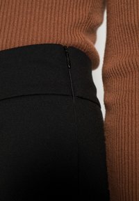 Musta korkeavyötäröinen housut, joissa on sileä pinta ja sivuvetoketju, yhdistetty ruskeaan ribbisaikkuun, jotka esittävät kontrastisia materiaaleja.