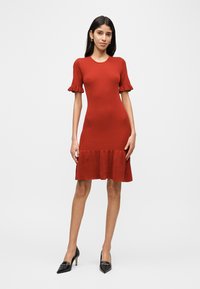 Φόρεμα από πλεκτό ύφασμα σε χρώμα σκουριάς με ριμπ ύφανση, κοντά μανίκια με βολάν και φαρδύ τελείωμα. Φορεμένο με μαύρα παπούτσια στιλέτο για αντίθεση.