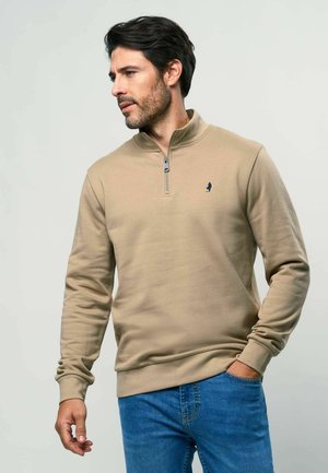 Smėlio spalvos medvilninis džemperis su ketvirčio užtrauktuku ant apykaklės, megztomis rankogaliais ir mažu mėlynu logotipu kairėje krūtinės pusėje. Dėvimas su mėlynais džinsais.