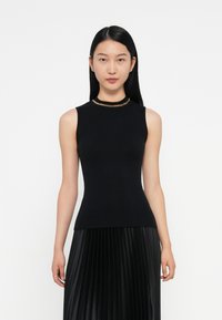 Top senza maniche nero con un design aderente e un accento di catena dorata sullo scollo. La modella indossa una gonna nera a pieghe.