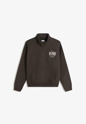 Bruine fleece trui met een kwart-ritssluiting bij de kraag, een voorvak en witte bedrukte tekst aan de bovenkant links. Zachte textuur met een ontspannen pasvorm.