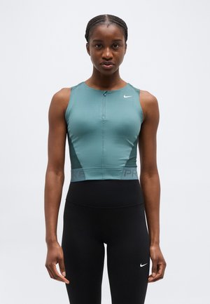 Atletična žena s ispletenom kosom, odjevena u tirkiznu majicu bez rukava sa zatvaračem i crne visokog struka tajice s Nike logotipima, stoji ispred bijele pozadine.