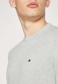 Giovane che indossa un maglione lavorato a maglia grigio chiaro con un piccolo logo rettangolare rosso, bianco e blu sul petto, su uno sfondo neutro.