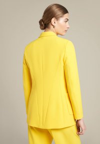 Żółta marynarka o dopasowanym kroju, z eleganckim designem, wyraźnie zarysowanymi ramionami i pojedynczym rozcięciem z tyłu. Posiada gładką teksturę oraz minimalistyczną kieszeń.