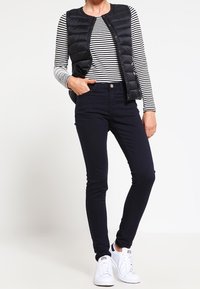 Kvinna i svart puffärmad väst, svart och vit randig långärmad tröja, svarta slim fit-byxor och vita sneakers står med ena handen i fickan.