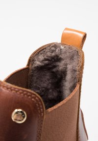 Botín de piel marrón con un exterior texturizado, suave forro de piel de color gris y una lengüeta de cuero. Decorado con un detalle de broche de latón.