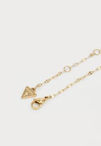 Gouden ketting met ovale schakels en een driehoekige hanger gegraveerd met "GUESS." Voorzien van een karabijnsluiting voor een veilige bevestiging.