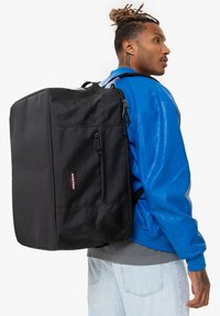 Černý batoh obdélníkového tvaru, vyrobený z odolné látky, se zipovými kapsami a nášivkou s logem. Nosený na zádech.