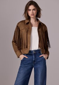 Brun mockaimo jacka med fransdetaljer, krage och dragkedja framtill, ihop med ljusblå vida jeans. En enkel vit topp under.