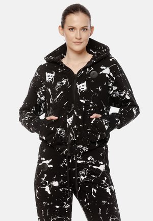 Kobieta ubrana w czarną bluzę z kapturem i spodnie z białym, abstrakcyjnym wzorem rozprysków farby, stojąca z rękami w kieszeniach bluzy.