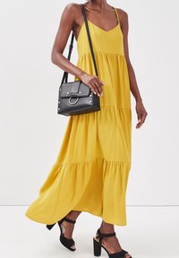 Robe longue jaune vif, avec des couches à volants, des bretelles spaghetti ajustables, accompagnée d'un petit sac à main noir et de sandales à talons carrés noires.