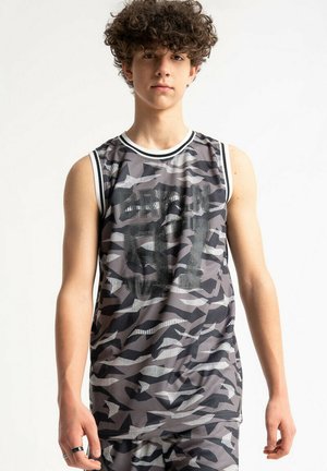 Nastolatek z kręconymi włosami, ubrany w szary i czarny bezrękawnik sportowy w kamuflażu z czarnymi literami i szorty na białym tle.