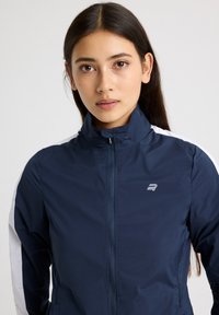 Giacca a vento navy con colletto alto, zip completa e accenti bianchi laterali. Presenta un logo bianco sul petto. Texture liscia.