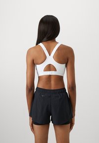 Kvinne med langt mørkt hår iført en hvit sports-bh med tykke stropper og svarte lagdelte treningsshorts, stående mot en ensfarget bakgrunn.