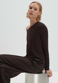 Braunes Langarmoberteil mit asymmetrischem Ausschnitt, kombiniert mit lockeren braunen Hosen. Glattes, weiches Material mit minimalen Designdetails.