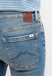 Jeans de mezclilla azul claro con un parche de cuero marrón del logo de Mustang y un bolsillo trasero con un detalle de costura en forma de montaña.