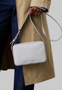 Borsa rettangolare bianca Calvin Klein con una superficie liscia, tracolla sottile e regolabile, e logo in rilievo sul davanti. Presenta una chiusura con zip.