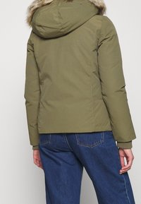 Person som bär en olivgrön vinterjacka med huva i faux päls och blå jeans, sedd bakifrån mot en enfärgad bakgrund.