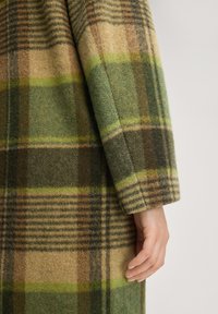 Wollmantel mit grünem, braunem und beige karriertem Muster. Lange Ärmel mit weicher Textur und detaillierter Nahtverarbeitung am Saum.