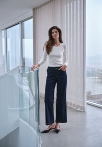 Donna con top bianco a maniche lunghe e jeans scuri a gamba larga in piedi accanto a una ringhiera in vetro in una stanza moderna con grandi finestre e tende trasparenti.