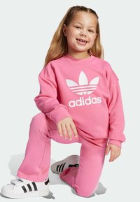 Ružová mikina adidas s bielym logom a zodpovedajúce ružové nohavice. Model nosí biele tenisky s čiernymi pruhmi. Mäkký, neformálny materiál a voľný strih.