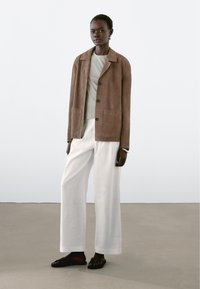 Braune Wildlederjacke mit vier vorderen Knöpfen, kombiniert mit einem hellgrauen T-Shirt, weißen Hosen mit weitem Bein und schwarzen Sandalen. Einfaches und sauberes Design.