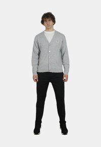 Cardigan grigio chiaro, abbottonato, con scollo a V, polsini e orlo a coste. Abbinato a pantaloni neri e sneakers. Modella in piedi con le mani in tasca.