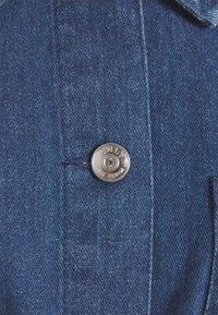 Bouton argenté portant l'étiquette "ONLY Blue Denim" sur un tissu en denim bleu avec des coutures visibles et une partie de poche.