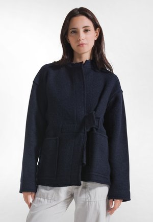 Donna con capelli scuri che indossa una giacca blu scuro con tasche frontali e cintura da annodare, abbinata a pantaloni chiari, in piedi su sfondo bianco.