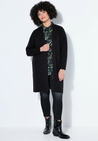 Femme aux cheveux bouclés portant un long cardigan noir, une chemise verte à motifs, un jean skinny noir et des bottines noires cloutées, debout contre un mur blanc.