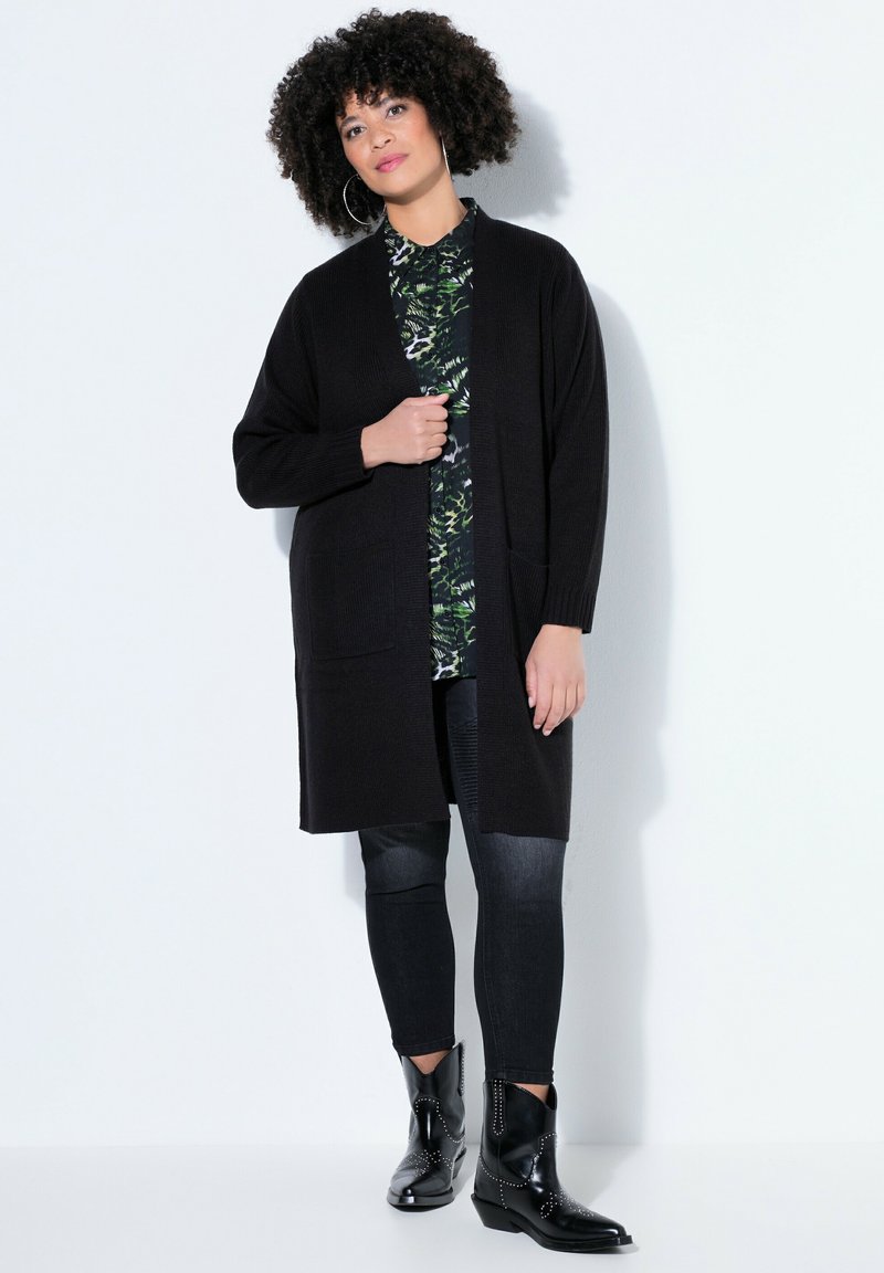 Femme aux cheveux bouclés portant un long cardigan noir, une chemise verte à motifs, un jean skinny noir et des bottines noires cloutées, debout contre un mur blanc.