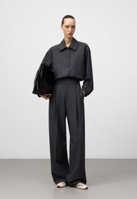 Donkergrijze oversized shirt met kraag en knoopsluiting aan de voorkant, gecombineerd met wijde broek. Model houdt een zwarte leren tas vast en draagt instapschoenen.