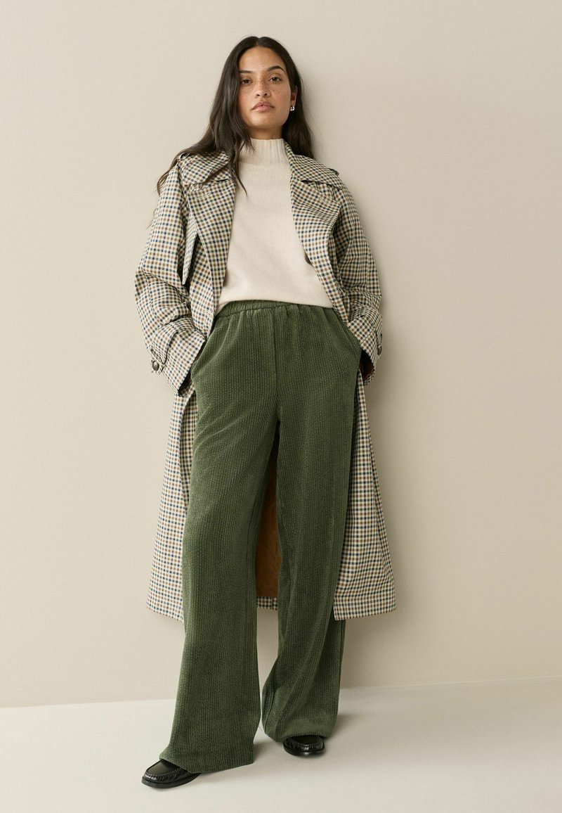 Long plaid coat in beige and green, paired with olive green corduroy wide-leg pants and a cream sweater. Glossy black shoes complete the look.