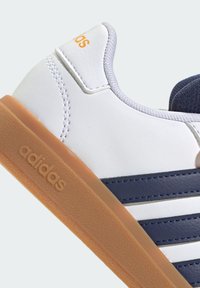 Zapatilla de cuero blanca con acentos en azul marino, tres rayas blancas y suela de goma. Presenta una textura suave y costuras en contraste.