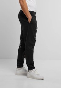 Black sweatpants with elastic waistband and cuffs. Features side pockets and a logo on the left thigh. Worn with white sneakers.