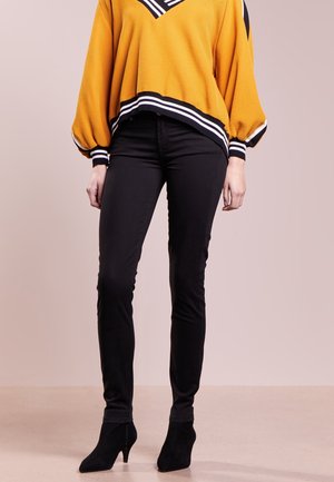 Femme portant un pantalon slim noir, des bottines noires à talons et un pull ample jaune moutarde avec une bordure rayée noire et blanche.