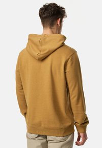 Bluza z kapturem w musztardowym kolorze, wykonana z mieszanki bawełny. Posiada ściągacze przy mankietach i u dołu, gładką teksturę oraz luźny krój. Pokazano widok z tyłu.