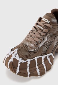 Pantof cu aspect sportiv de culoare maronie, cu accente din plasă, talpă din cauciuc texturat și detalii albe asemănătoare cu mărgele în zona degetelor. Pănjeri și logo-ul mărcii sunt vizibile.