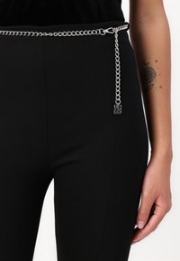 El cinturón de cadena plateada presenta un clip rectangular, un detalle de cadena colgante y un charm con el logotipo en negro, llevado sobre una prenda negra ajustada.