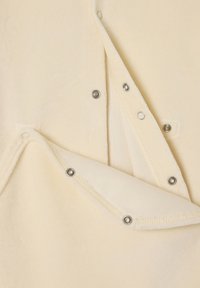 Cremefarbenes Kleidungsstück mit weicher Textur, das über Druckknöpfe an der Vorderseite verfügt und am unteren Rand eine geschichtete, gefaltete Gestaltung aufweist.
