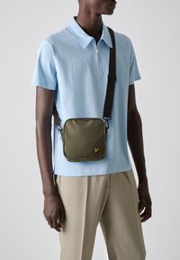 Geantă crossbody de culoare verde măslinie, confecționată dintr-un material durabil, cu formă pătrată, cu închidere cu fermoar, prezentând un emblema galbenă. Purtați deasupra unei cămăși polo de culoare albastru deschis.